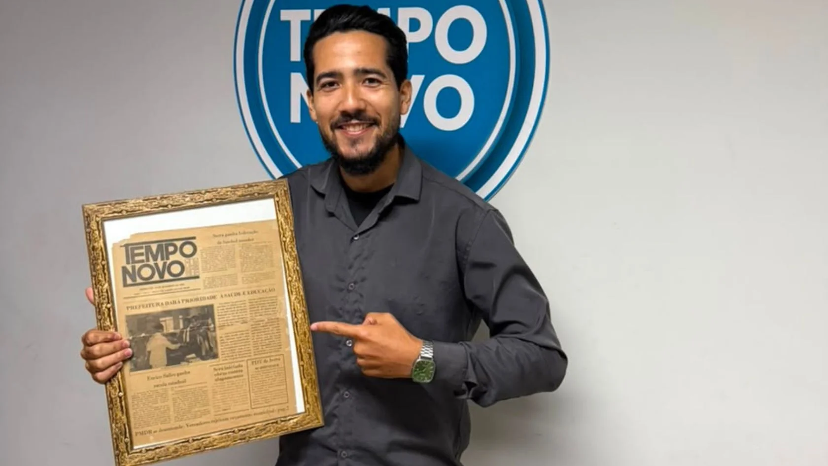 Wal Júnior, novo repórter do Tempo Novo, sorri ao lado de edição histórica emoldurada do jornal.