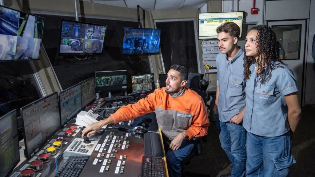 Profissional opera painel de controle industrial enquanto dois jovens observam, em imagem sobre vagas de estágio na Serra