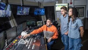 Profissional opera painel de controle industrial enquanto dois jovens observam, em imagem sobre vagas de estágio na Serra