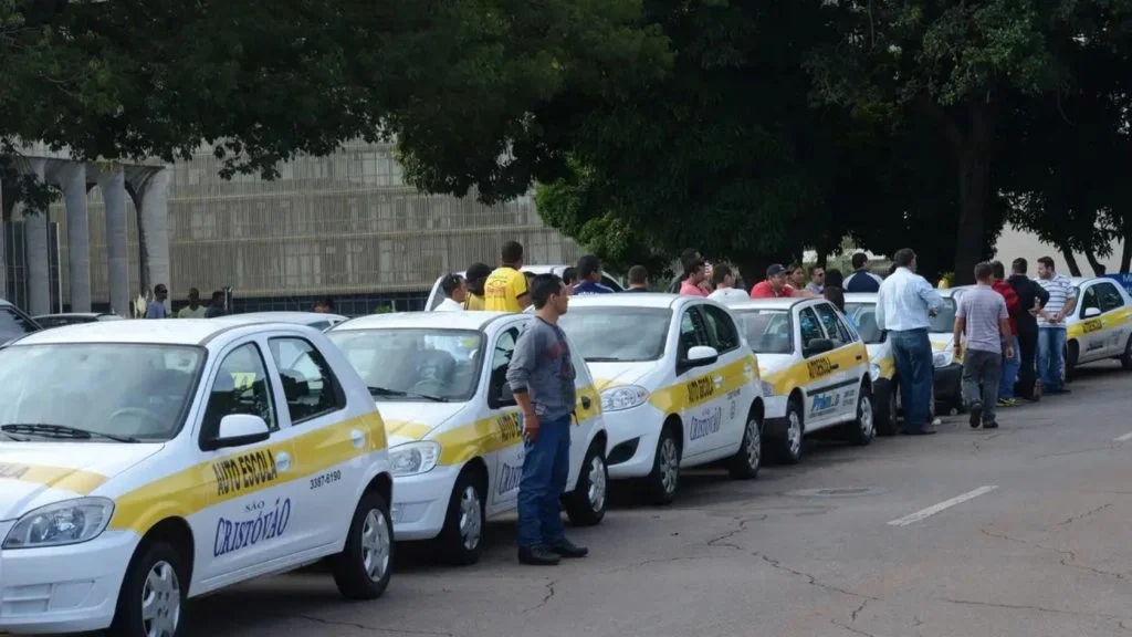 Carros de autoescola enfileirados em rua com candidatos ao lado, cenário afetado pelo exame de direção com carro particular
