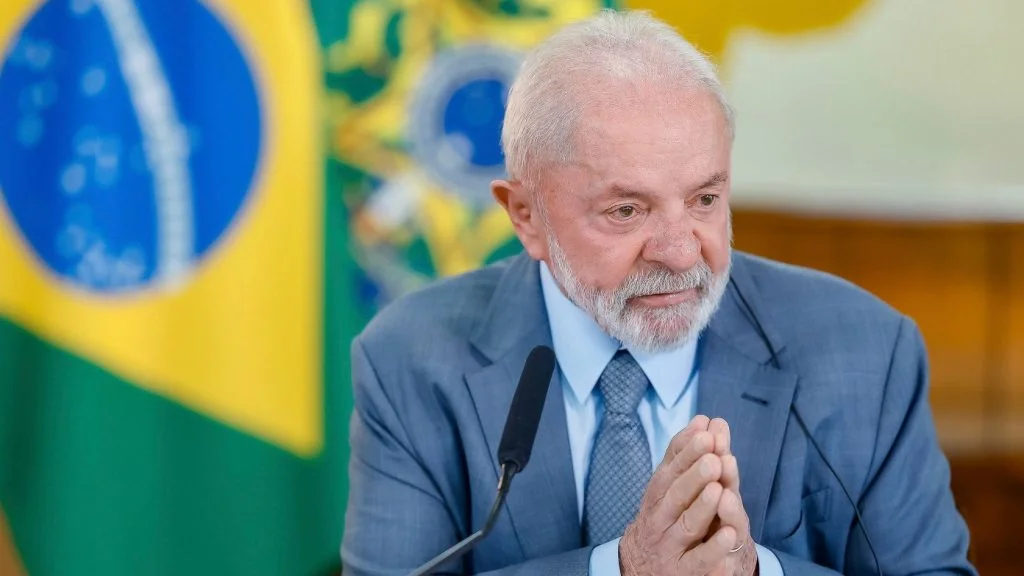 Presidente Lula, de terno azul e mãos juntas, diante da bandeira do Brasil; ele prevê o fim da escala 6x1 ainda este ano