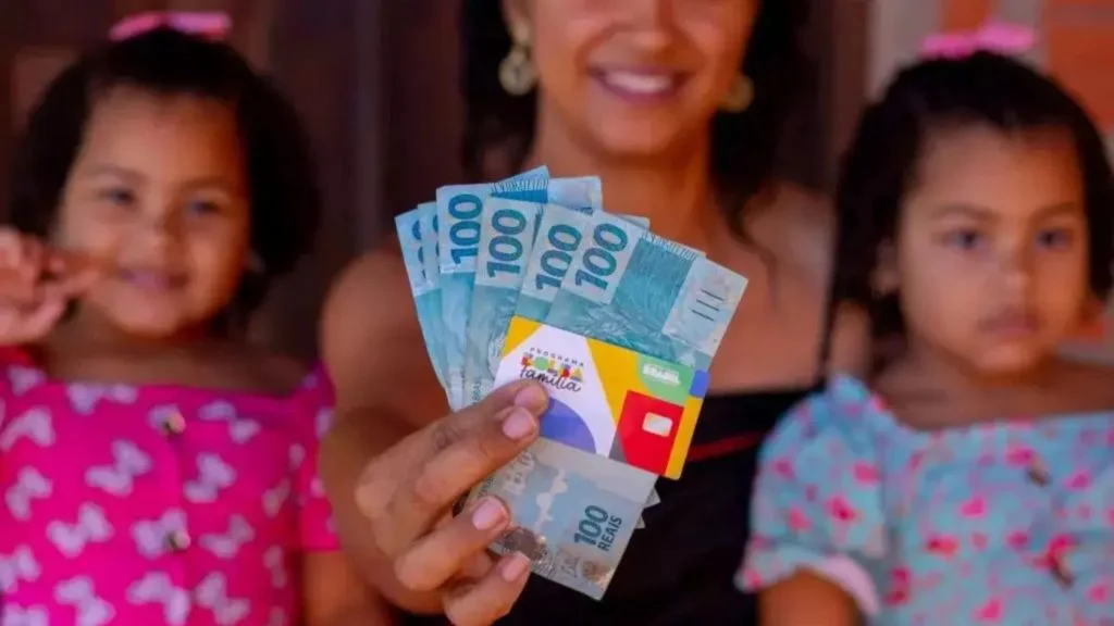 Mulher segura notas de R$ 100 e cartão do Bolsa Família ao lado das filhas