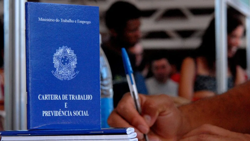 Carteira de Trabalho em destaque para a feira de empregos na Serra, com mão segurando caneta e pessoas ao fundo