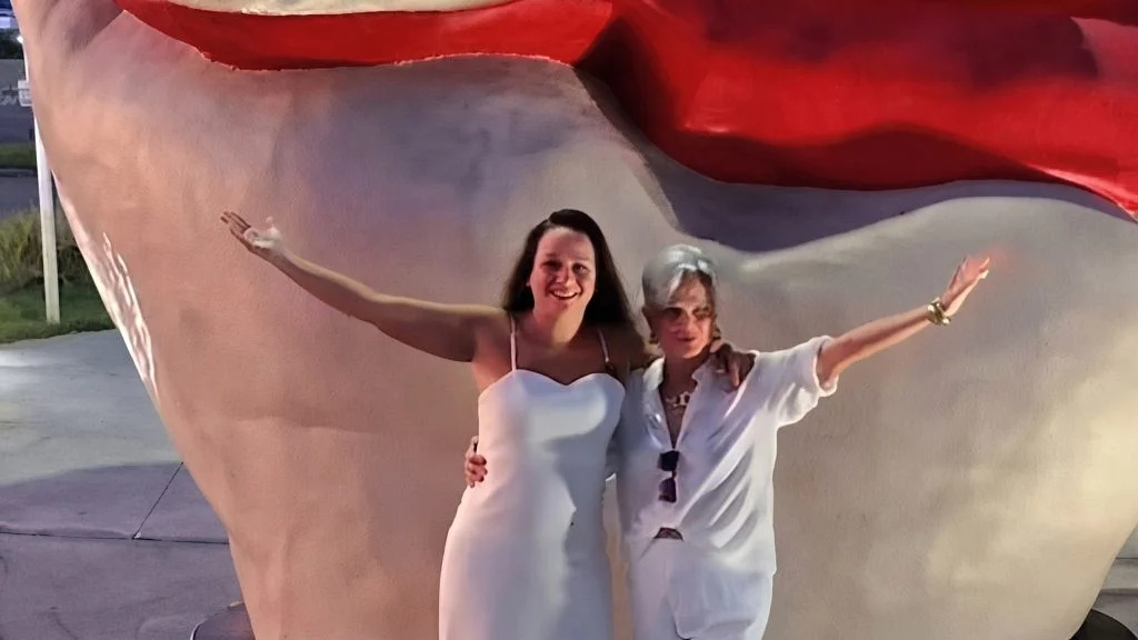 Penélope Feu Rosa sorri ao lado de outra mulher, os dois de braços abertos diante de escultura com a bandeira do Brasil