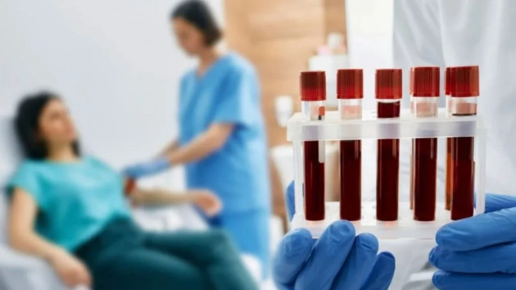 Mãos com luvas seguram tubos de exame de sangue; ao fundo, profissional de saúde faz coleta em paciente