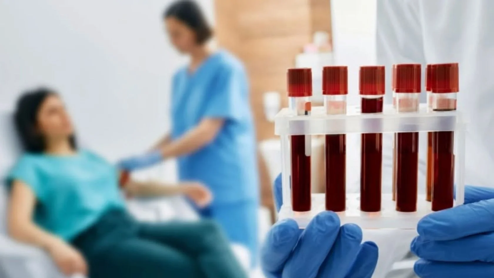 Mãos com luvas seguram tubos de exame de sangue; ao fundo, profissional de saúde faz coleta em paciente