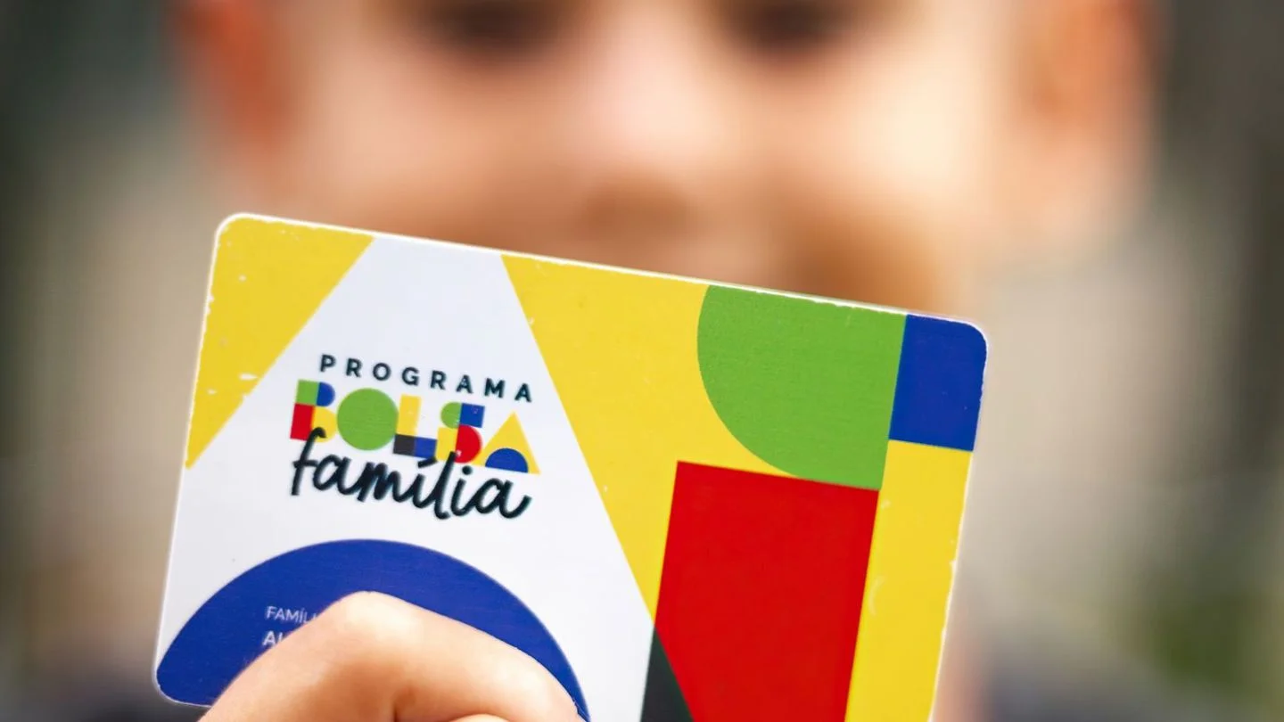 Criança segura cartão do Bolsa Família em primeiro plano; pagamento de outubro chega com reajuste