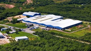Vista aérea de fábrica da Deca, com galpões brancos e azuis cercados por área verde e caminhões no pátio