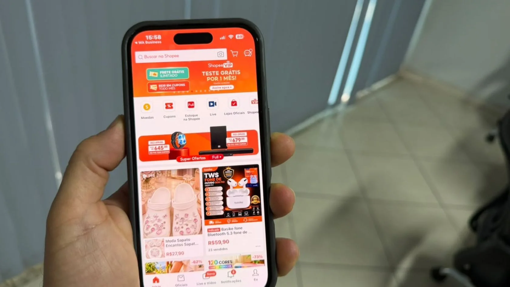 Mão segura celular com a tela inicial do app da Shopee, onde ficam as moedas usadas na promoção da Shopee
