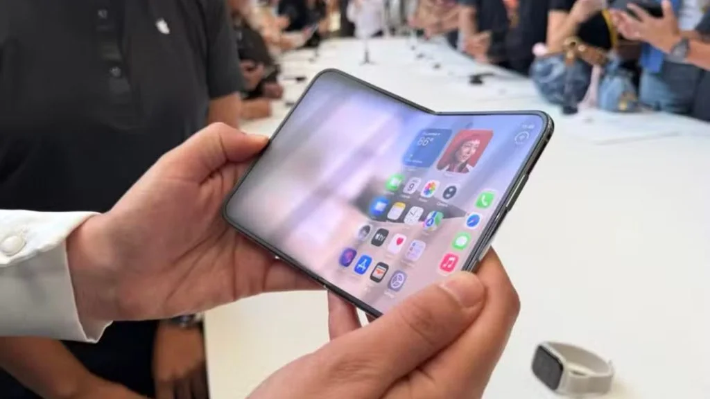 Mãos seguram o iPhone dobrável parcialmente aberto, com a tela interna exibindo aplicativos, em área de demonstração