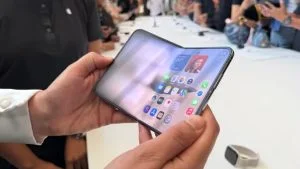 Mãos seguram o iPhone dobrável parcialmente aberto, com a tela interna exibindo aplicativos, em área de demonstração