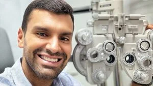 Serginho Vidigal sorri ao lado de um refrator, aparelho de exame de vista, em consultório médico