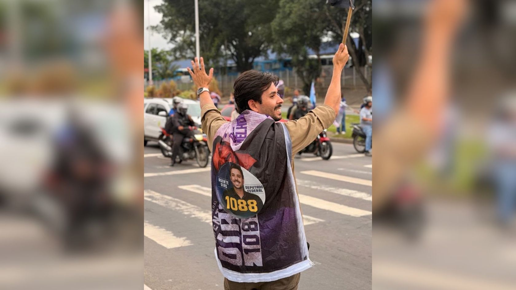 Vereador Agente Dias, com bandeira da campanha a deputado federal, acena com os braços erguidos em avenida