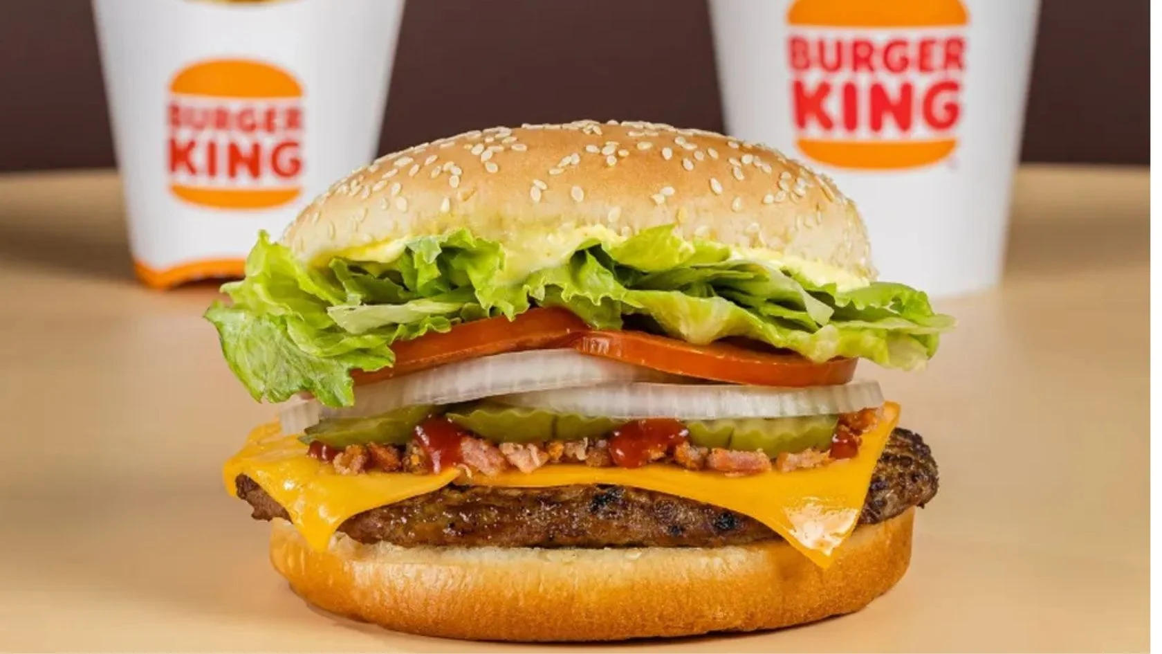 Hambúrguer com queijo, bacon e salada em frente a copos do Burger King; rede dará lanche grátis nesta sexta
