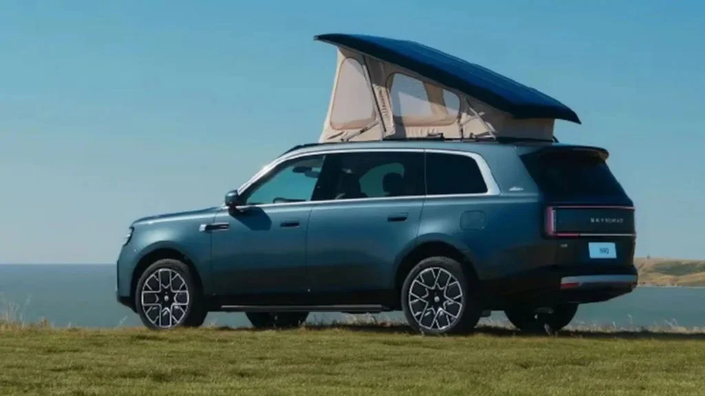 Carro da Xiaomi SkyNomad N90 azul com teto elevado e tenda bege, estacionado em gramado com o mar ao fundo