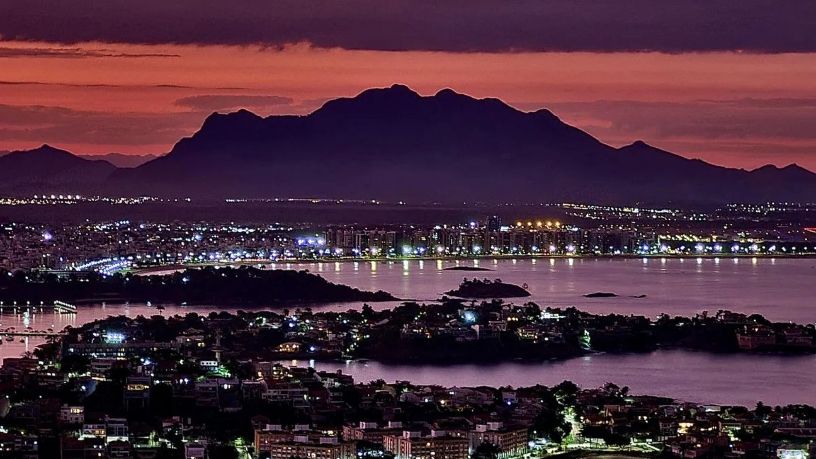 Vista aérea ao anoitecer, com baía, ilhas, prédios iluminados e serra ao fundo, ilustra matéria sobre preço do apartamento na Serra